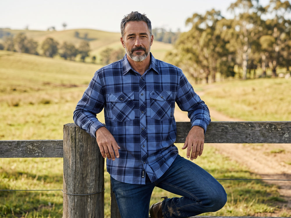 Man wearing a blue plaid shirt leaning against a wooden fence in a rural setting.