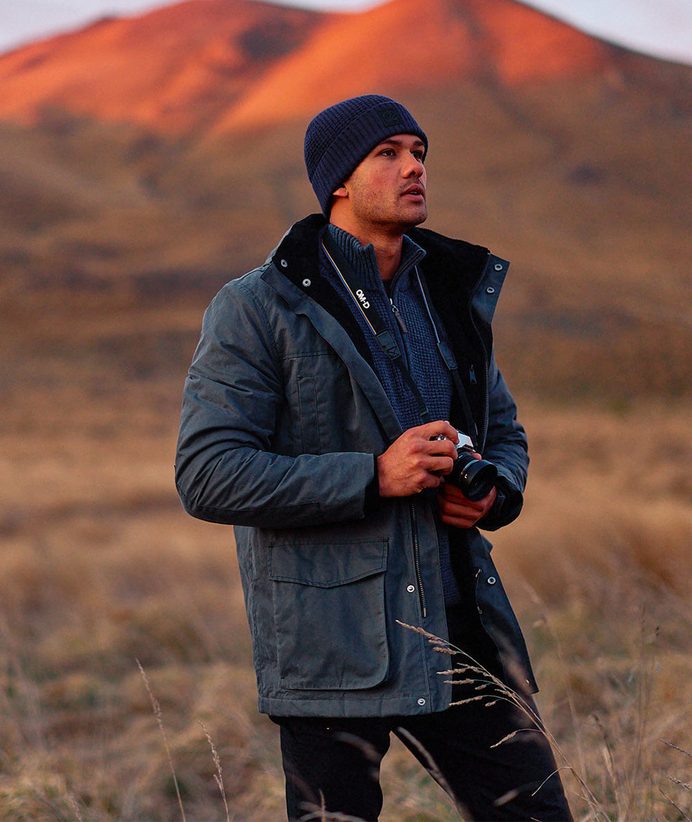 Man in a dark jacket and beanie standing in a field with mountains in the background