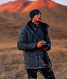 Man in a dark jacket and beanie standing in a field with mountains in the background