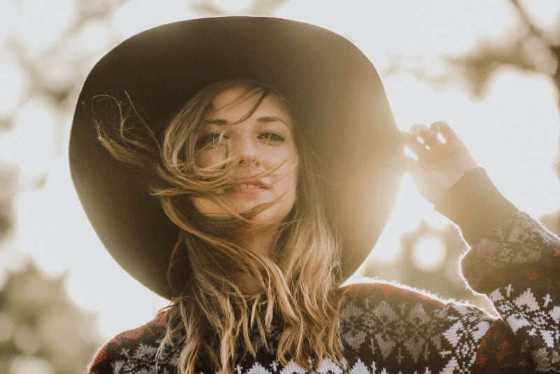 Female farmer wearing a wide-brimmed hat and patterned jumper outdoors with the sun shining behind her.