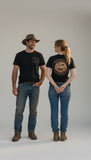 Male and female models wearing the black Outback Clothing Co OCC Muster Tee showing the front chest logo and large back artwork.