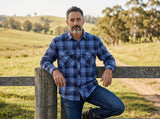 Man wearing a blue plaid shirt leaning against a wooden fence in a rural setting.