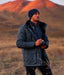 Man in a dark jacket and beanie standing in a field with mountains in the background