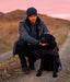 Man wearing Newton Navy Blue Beanie in outdoor setting with a black dog, both on a path with a pink sky behind them.