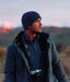 Man wearing a blue beanie and jacket with a camera around his neck, standing in a scenic outdoor setting.