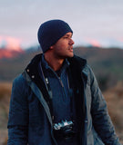 Man wearing a blue beanie and jacket with a camera around his neck, standing in a scenic outdoor setting.