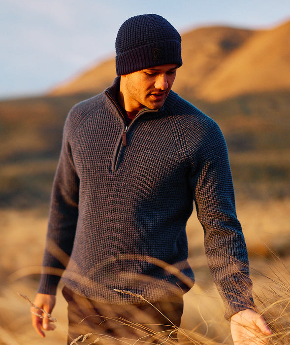 Man wearing a dark sweater and Newton Navy Blue knit beanie standing in a field with mountains in the background.